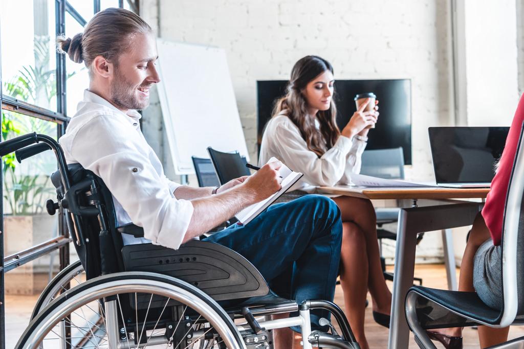 Lächelnder behinderter Geschäftsmann im Rollstuhl macht sich am Arbeitsplatz Notizen - Foto, Bild