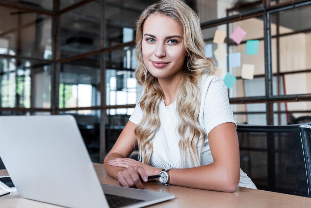 bela jovem empresária usando laptop e sorrindo para a câmera no escritório
 - Foto, Imagem