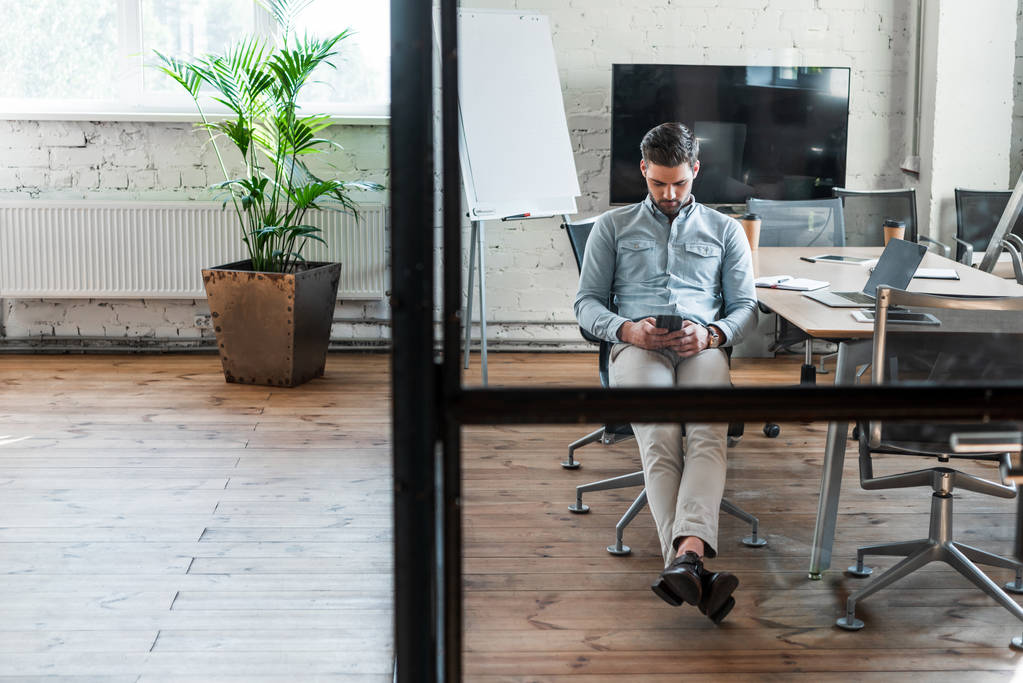 bonito jovem empresário usando smartphone enquanto sentado no local de trabalho
 - Foto, Imagem