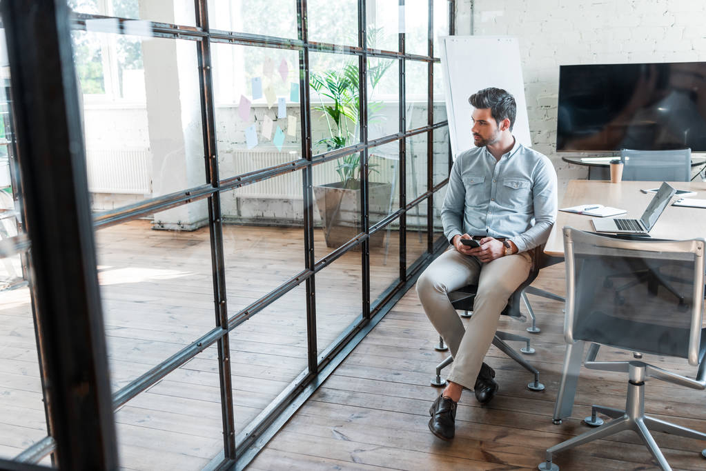 visão de alto ângulo do jovem empresário usando smartphone e olhando para longe enquanto sentado no escritório
 - Foto, Imagem