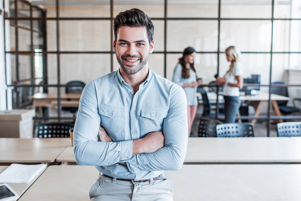 handsome young businessman with crossed arms smiling at camera in office - Photo, Image