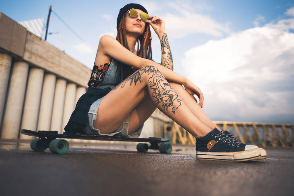 fiatal lány tetoválás és a kék sapka raszta ül a longboard, a háttérben a betonszerkezet - Fotó, kép