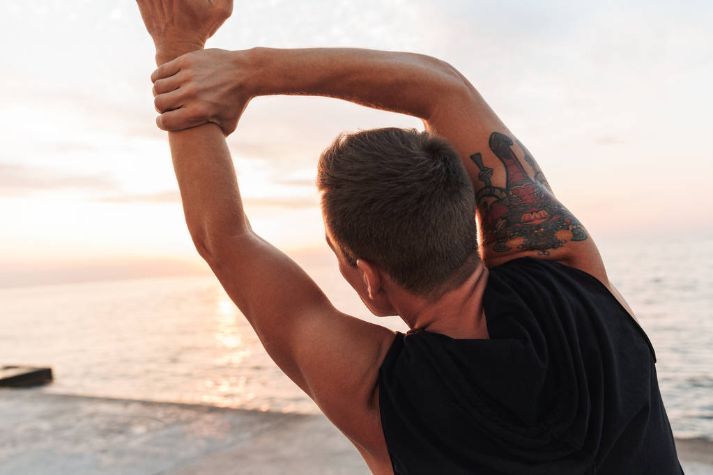Vista posterior de la imagen de joven deportista fuerte guapo al aire libre en la playa hacer ejercicios de estiramiento
. - Foto, Imagen