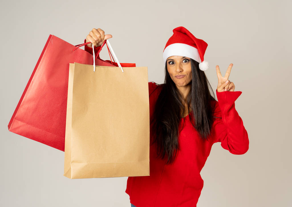 Mujer joven lista para Navidad con bolsas de compras de papel en rojo y sombrero de Santa Claus fondo gris aislado en estar listo para la Navidad regalos celebración consumismo y concepto de ventas
. - Foto, Imagen