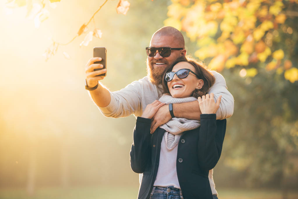 Joven feliz abrazo sonriente pareja (hombre y mujer) con teléfono haciendo selfie mientras camina en el jardín de otoño contra amarillo-verde fondo soleado (puesta de sol
) - Foto, Imagen