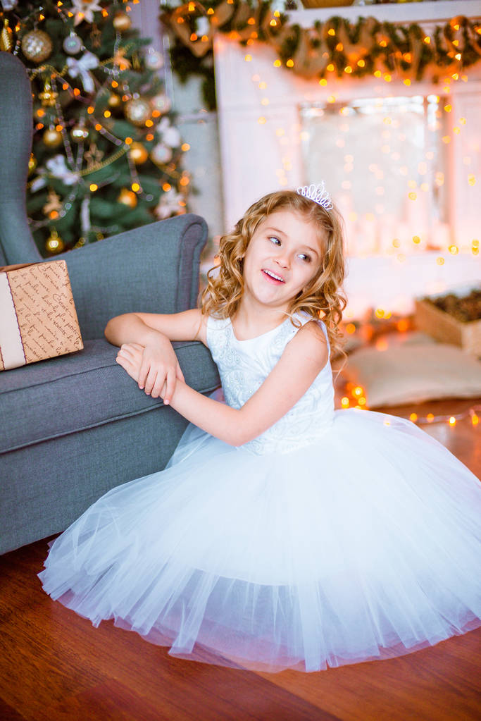 niedliches kleines Mädchen mit lockigem blondem Haar zu Hause in der Nähe eines Weihnachtsbaums mit Geschenken und Girlanden und einem dekorierten Kamin  - Foto, Bild