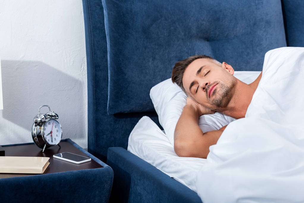 Erwachsener Mann schläft im Bett neben Wecker und Smartphone mit leerem Bildschirm zu Hause - Foto, Bild