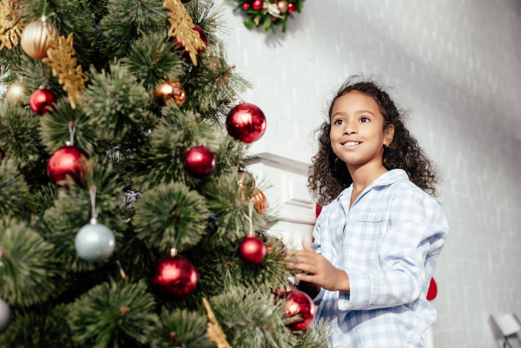 smiling adorable african american child in pajamas decorating christmas tree with baubles and looking away at home - Photo, Image