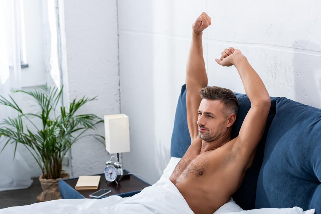 selective focus of shirtless man with raised arms doing stretch in bed at home - Photo, Image