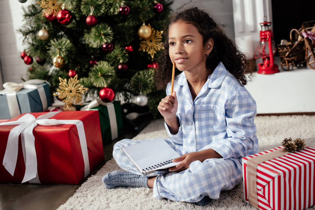 adorable niño afroamericano pensativo en pijama sentado con copybook cerca del árbol de Navidad en casa
 - Foto, imagen