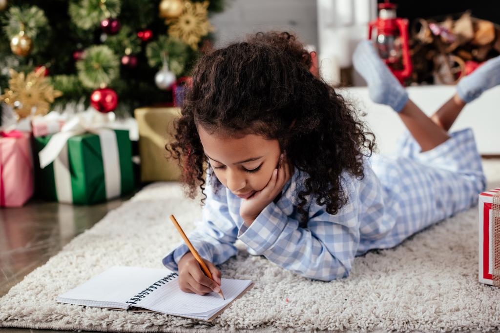 adorable Afryki amerykański dziecko w piżamie leży na podłodze i coś pisze do rezerwowym w domu christmas koncepcji - Zdjęcie, obraz