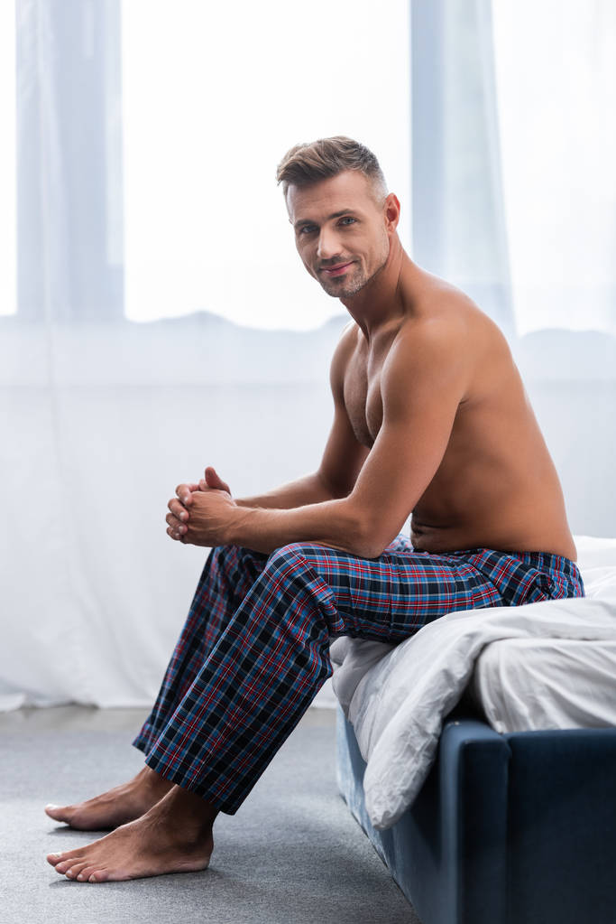 smiling shirtless adult man sitting on bed during morning time at home - Photo, Image