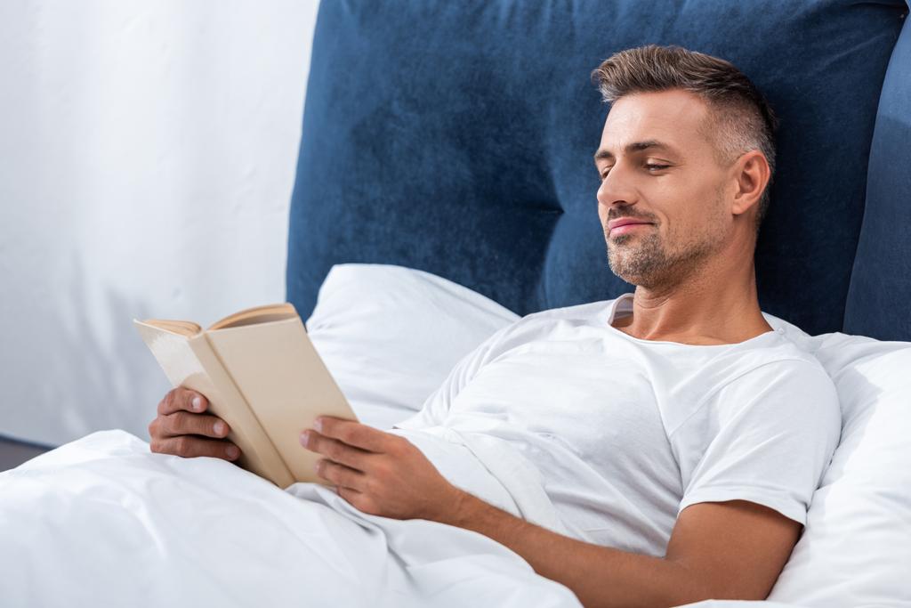 sonriente hombre leyendo libro mientras está acostado en la cama durante la mañana en casa
 - Foto, Imagen