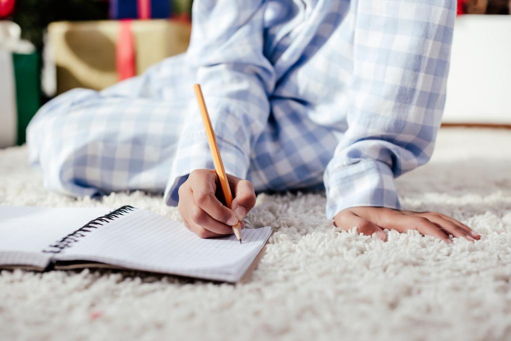 abgeschnittenes Bild eines afrikanisch-amerikanischen Kindes im Pyjama, das zu Hause mit Bleistift etwas in Notizbuch schreibt - Foto, Bild