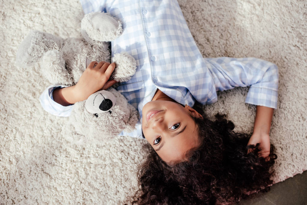テディベアの部屋でカーペットの上で横になっていると、カメラ目線はパジャマ姿で愛らしいアフリカ系アメリカ人子供の平面図 - 写真・画像