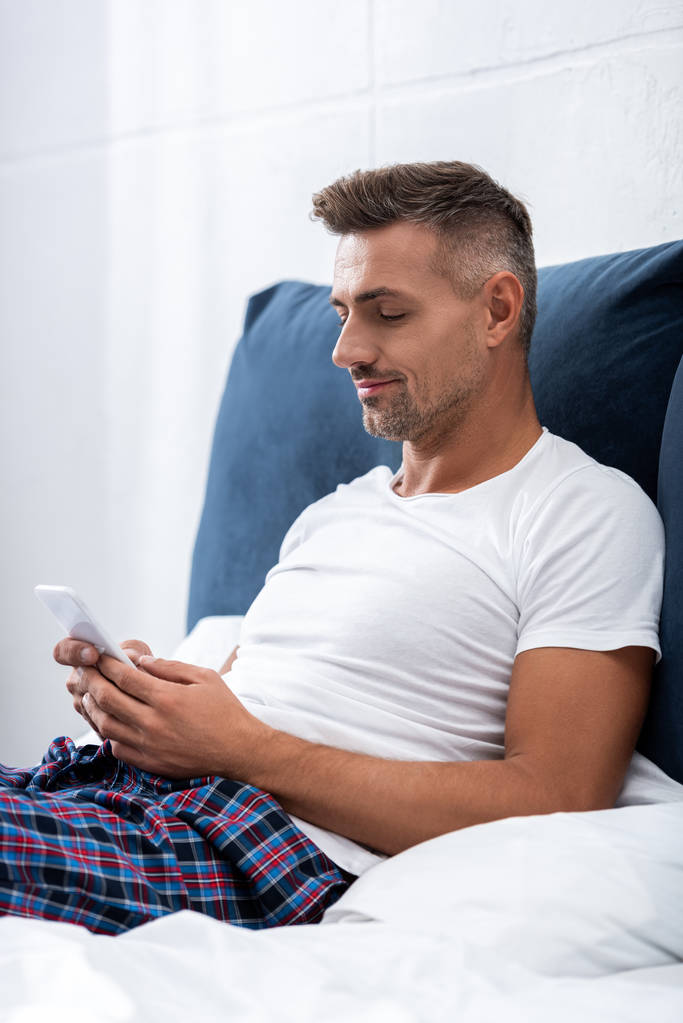 чоловік у білій футболці використовує смартфон під час ранкового часу в ліжку вдома
 - Фото, зображення