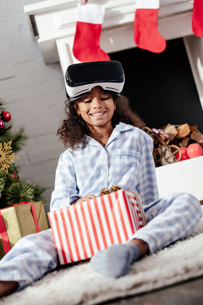 χαμογελώντας αξιολάτρευτο παιδί αφρικανική αμερικανική πιτζάμες και σετ κεφαλής εικονικής πραγματικότητας που βλέπουν στο σπίτι δώρο Χριστουγέννων - Φωτογραφία, εικόνα