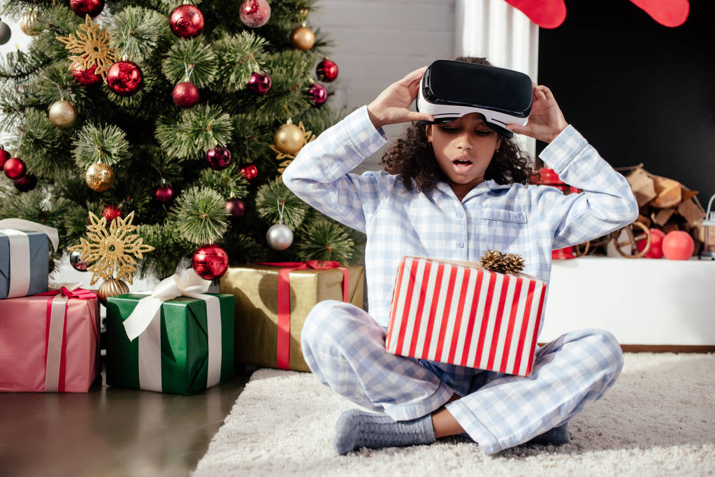 yllättynyt suloinen afrikkalainen amerikkalainen lapsi pyjamassa ja virtuaalitodellisuuskuulokkeissa katselemassa joululahjaa kotona
 - Valokuva, kuva