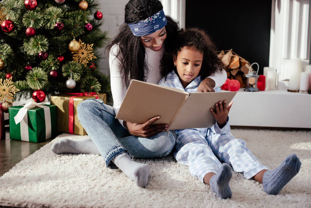 african american mother and daughter reading book together near christmas tree at home - Photo, Image