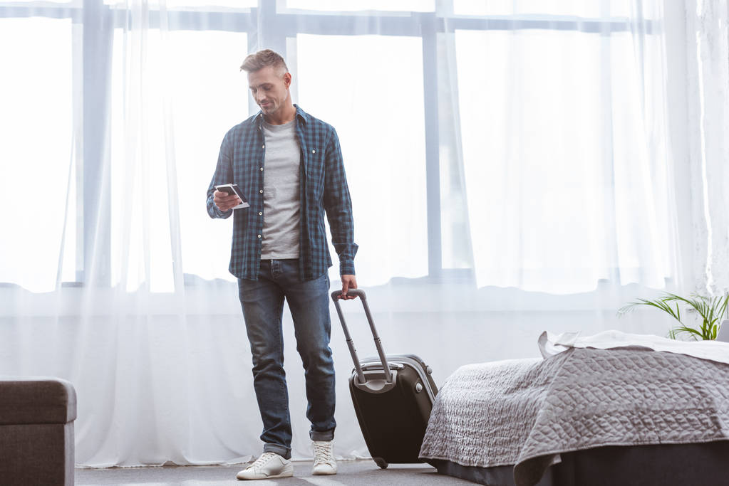 vidám ember használ smartphone és szállító kerekes táska otthon  - Fotó, kép