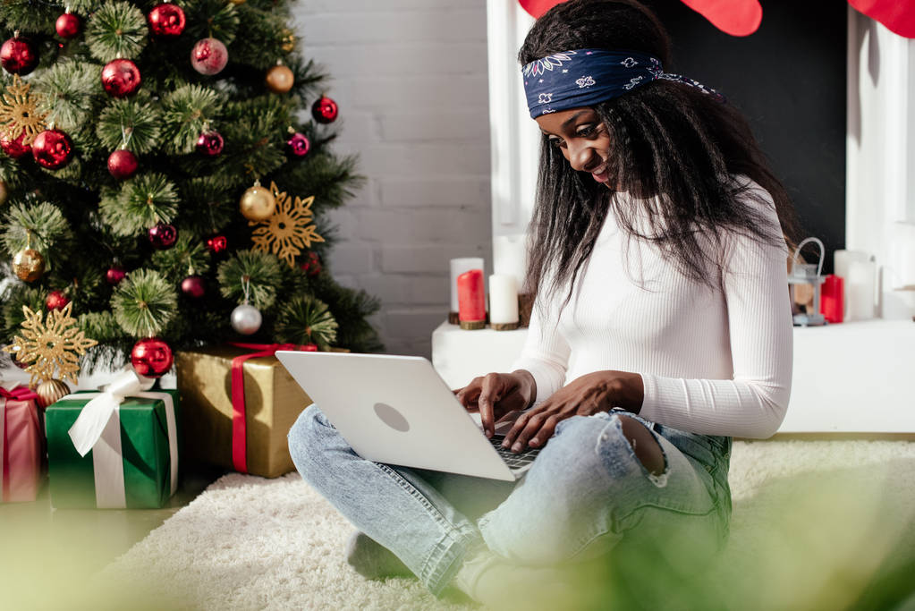 sonriendo atractiva mujer afroamericana utilizando portátil en el suelo en casa, concepto de Navidad
 - Foto, imagen
