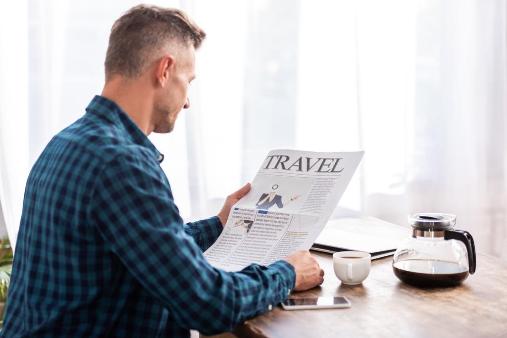 vista trasera del hombre sentado a la mesa y leyendo el periódico de viaje en la cocina en casa
 - Foto, Imagen