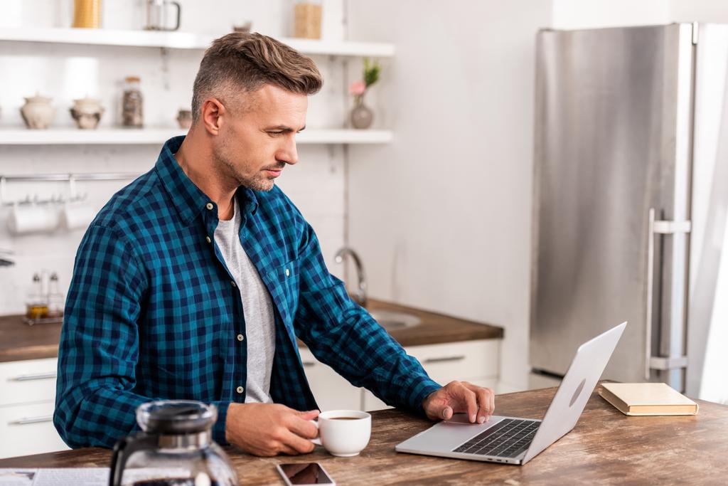 όμορφος άνδρας στο καρώ πουκάμισο κρατώντας το φλιτζάνι του καφέ και να χρησιμοποιούν φορητό υπολογιστή στο σπίτι - Φωτογραφία, εικόνα