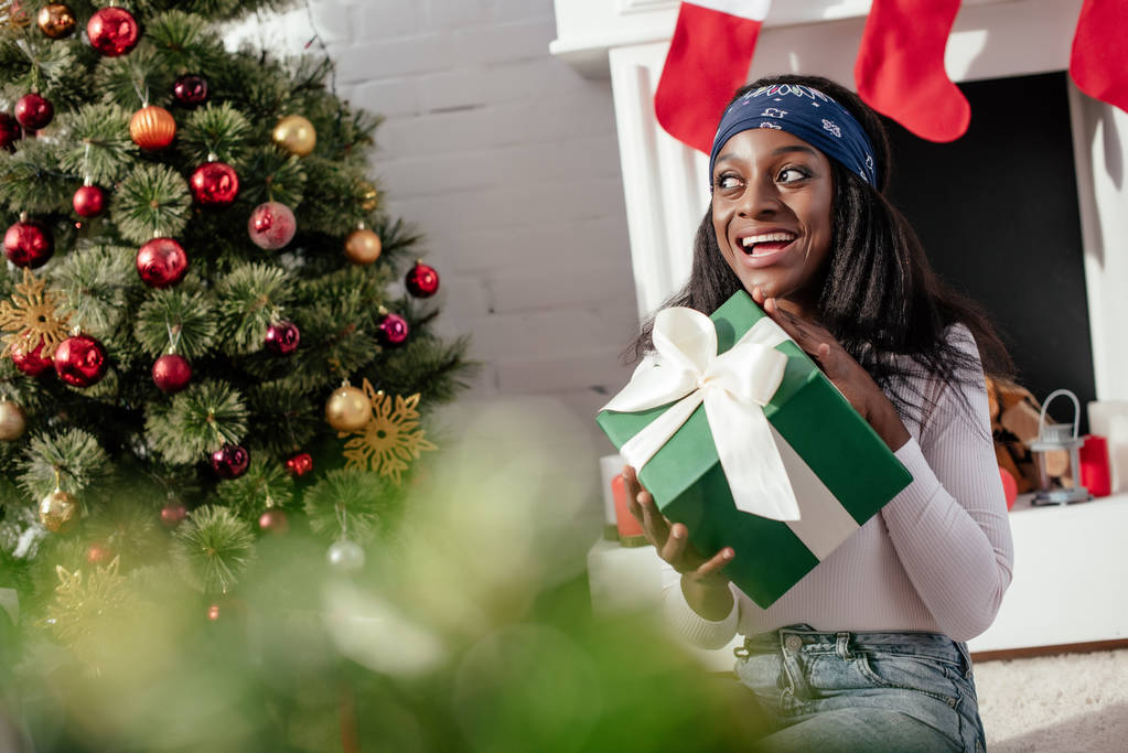 excited attractive african american woman holding christmas present at home and looking away - Photo, Image