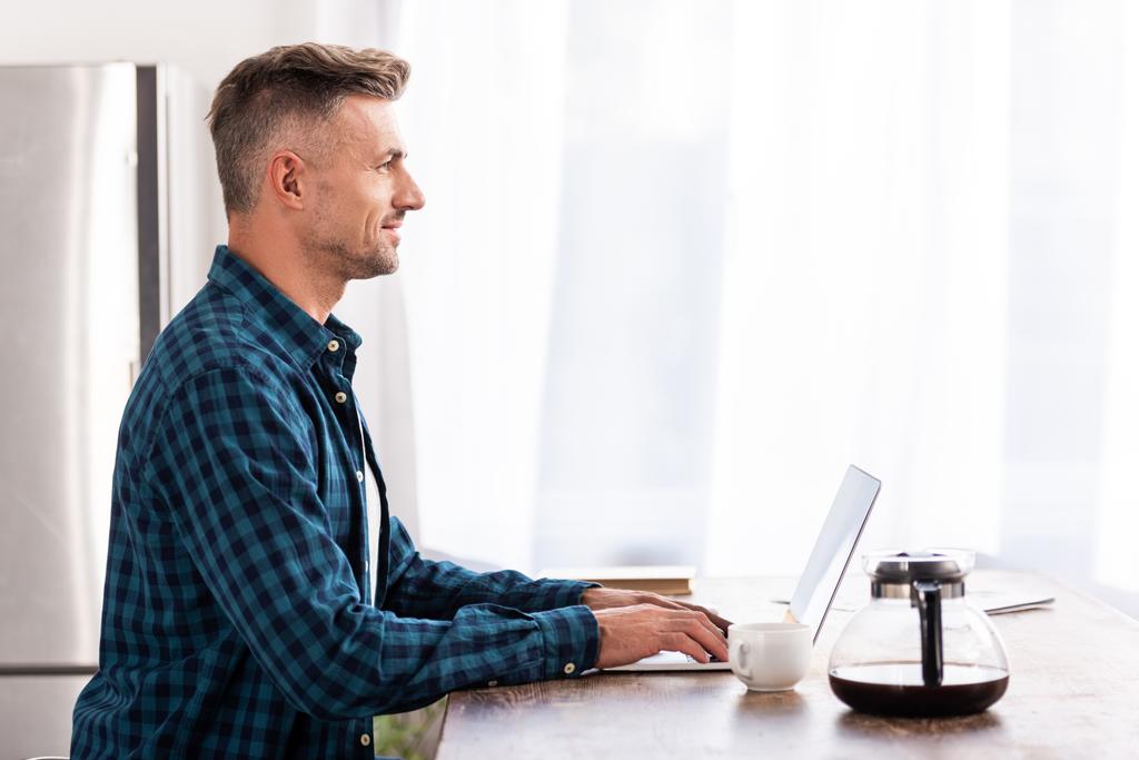 vista lateral do homem sorridente em camisa quadriculada usando laptop em casa
 - Foto, Imagem