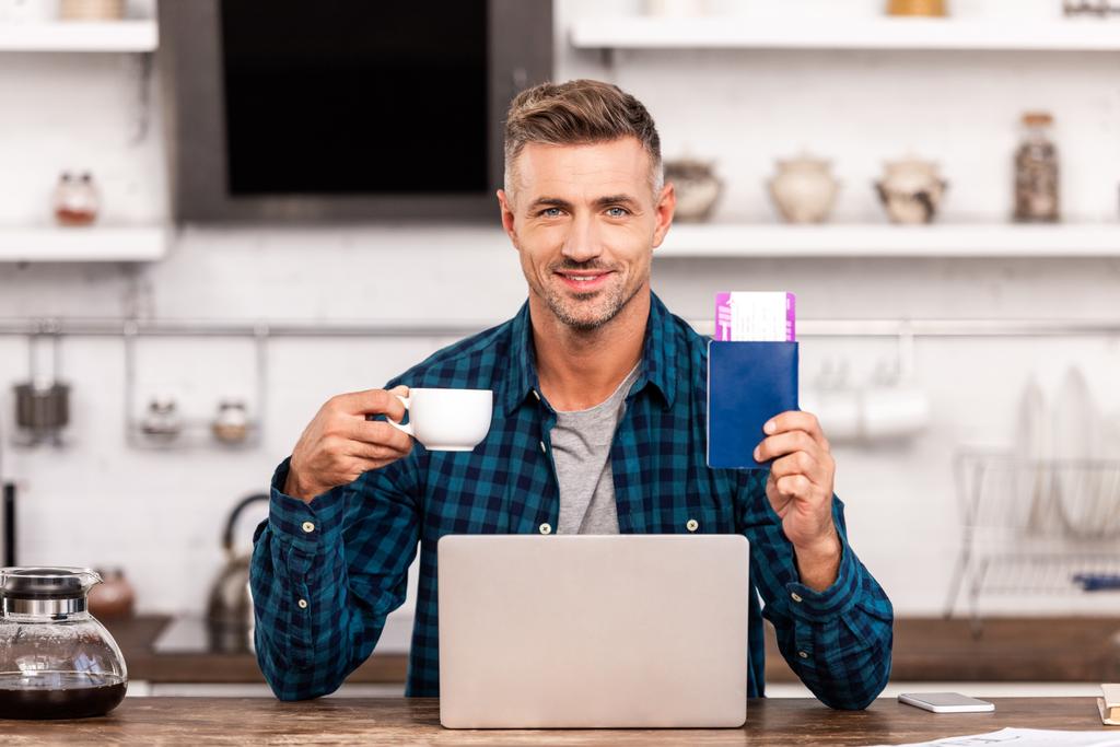 hombre feliz sosteniendo la taza de café y pasaporte con billete mientras usa el ordenador portátil en casa
  - Foto, imagen