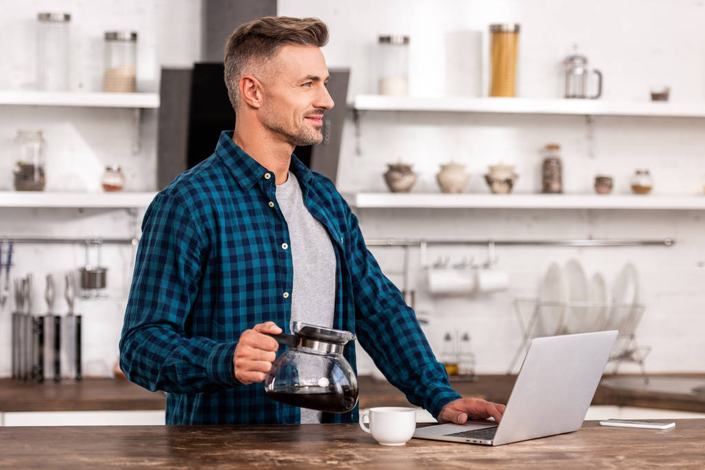 bonito sorrindo homem segurando panela de café e usando laptop em casa
 - Foto, Imagem