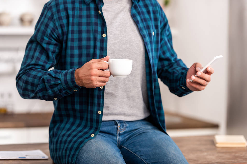 plan recadré de l'homme en chemise à carreaux tenant tasse de café et en utilisant smartphone à la maison
  - Photo, image