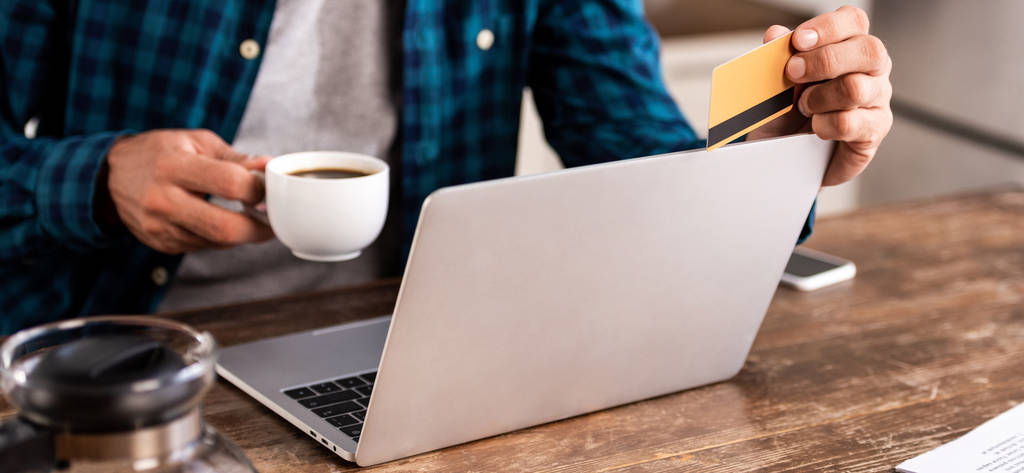 oříznutý snímek člověka drží šálek kávy a kreditní karty při používání notebooku doma - Fotografie, Obrázek