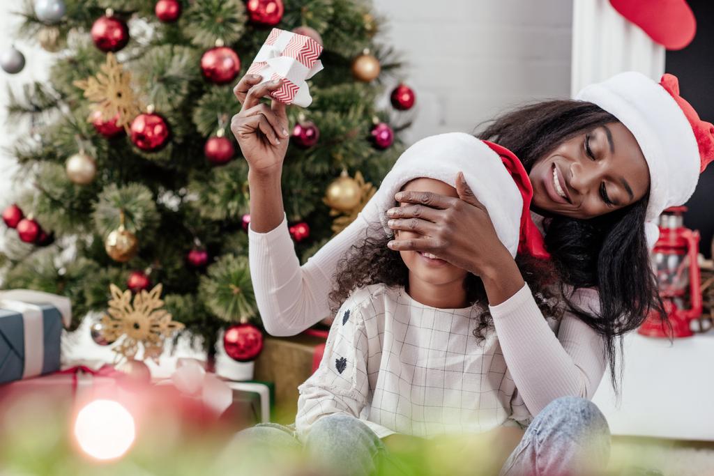 улыбающаяся африканская американская мать в шляпе Санта-Клауса, удивительная дочь с рождественским подарком дома
 - Фото, изображение