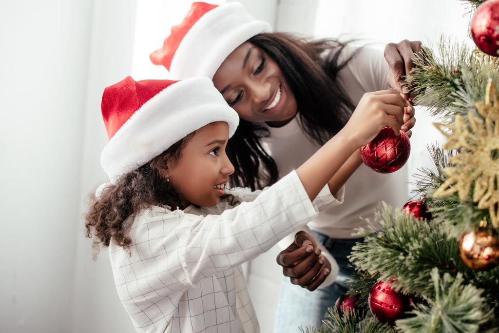 african american mother and daughter in santa claus hats decorating christmass tree together at home - Photo, Image