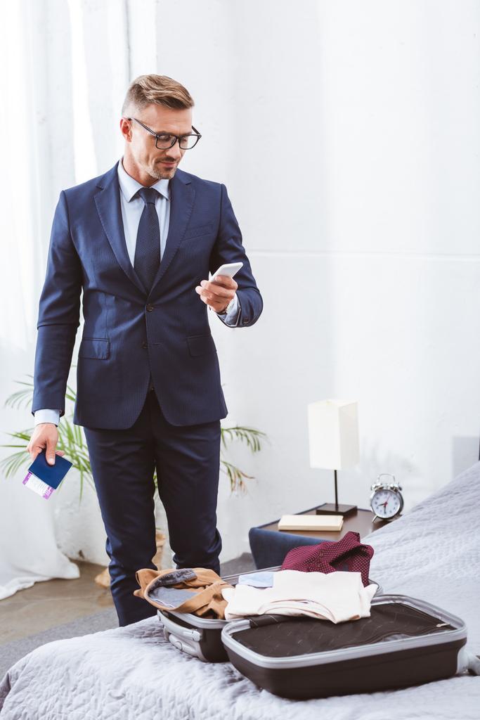 smiling businessman in suit and eyeglasses holding passport with boarding pass and using smartphone while packing suitcase at home - Photo, Image