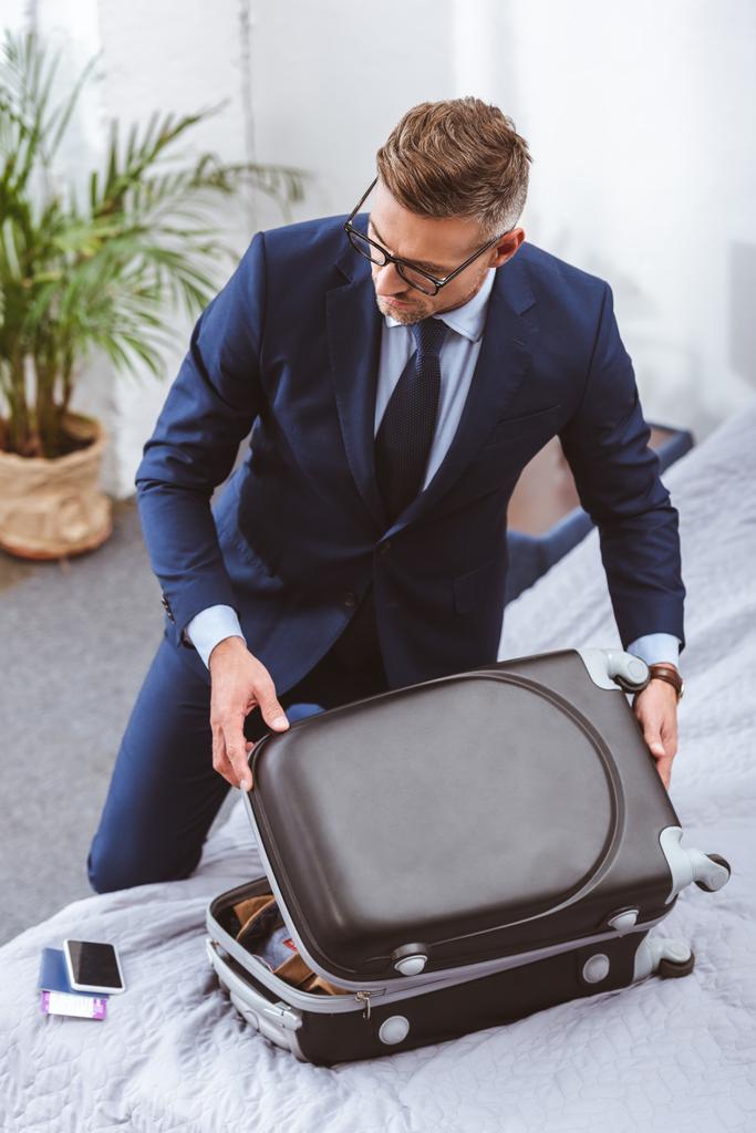 Blick aus der Vogelperspektive: Geschäftsmann in Anzug und Brille packt Koffer zu Hause  - Foto, Bild