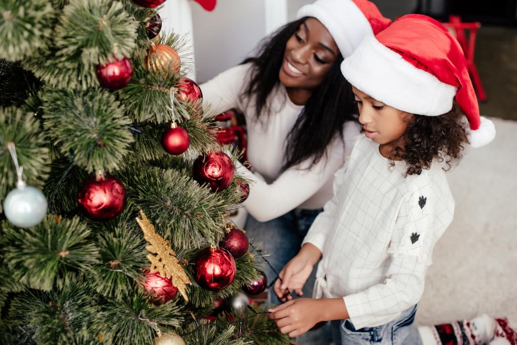 african american mother and daughter in santa claus hats decorating christmass tree together at home - Photo, Image