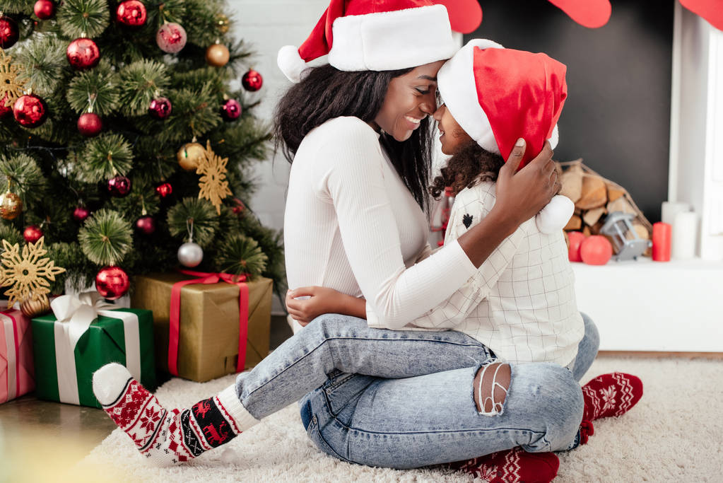 szczęśliwy african american kobieta i córka w santa claus kapelusze przytulanie w urządzonych pokojach na Boże Narodzenie w domu - Zdjęcie, obraz
