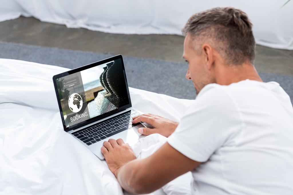 man in white t-shirt using laptop with booking website on bed - Photo, Image
