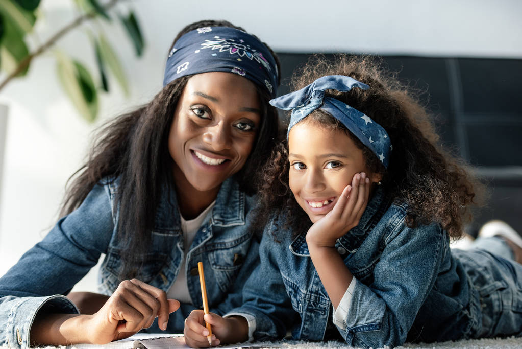 Ať se usmívám africká americká žena a dcera zároveň dělat domácí úkoly doma při pohledu na fotoaparát - Fotografie, Obrázek