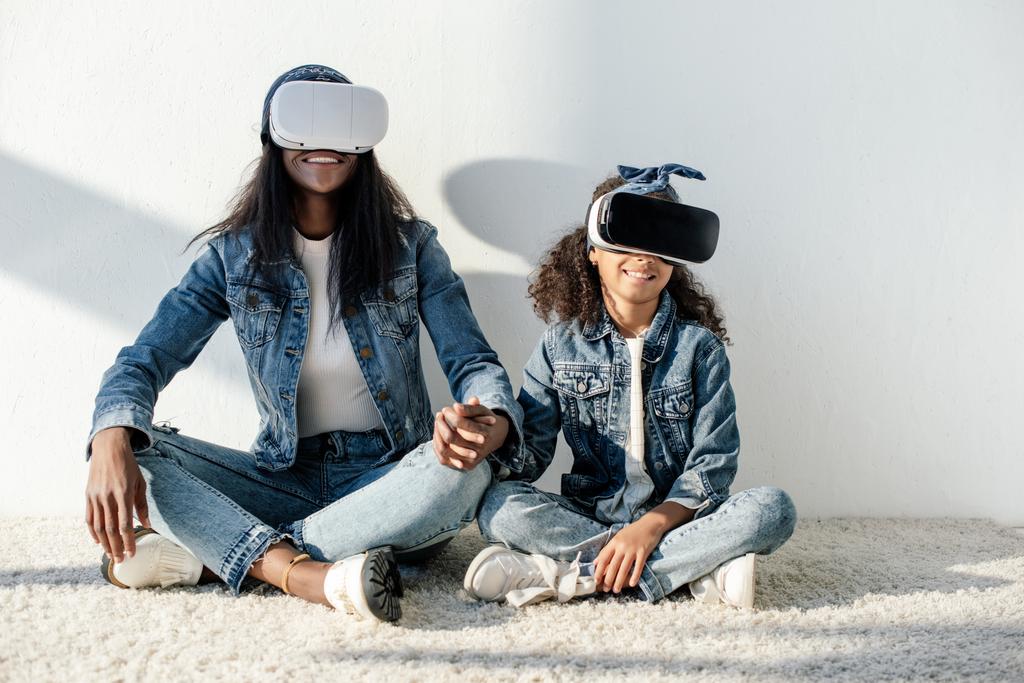 african american mother and daughter in similar clothing and vr headsets holding hands while sitting on floor at home - Photo, Image