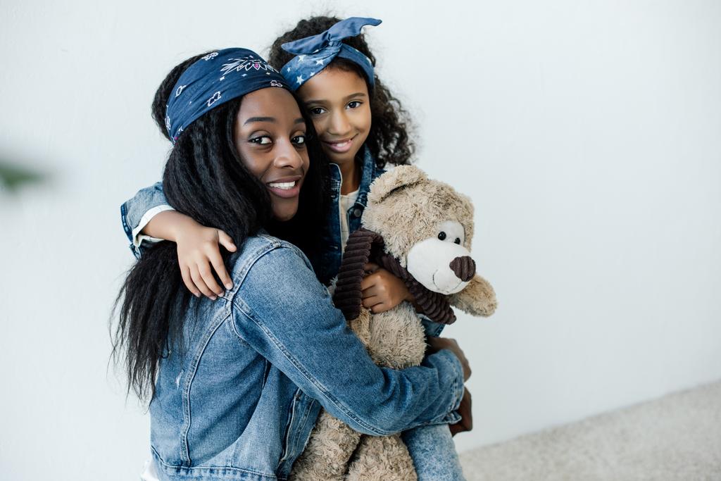retrato de la sonriente hija afroamericana con oso de peluche abrazando a la madre en casa
 - Foto, Imagen