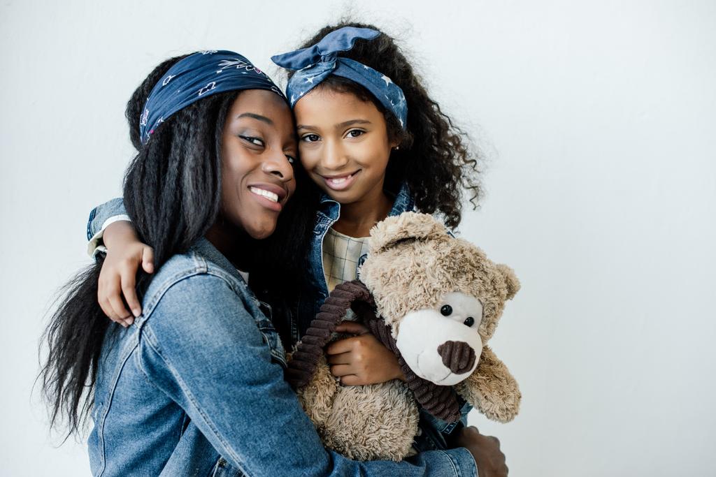 portrait of smiling african american daughter with teddy bear hugging mother at home - Photo, Image