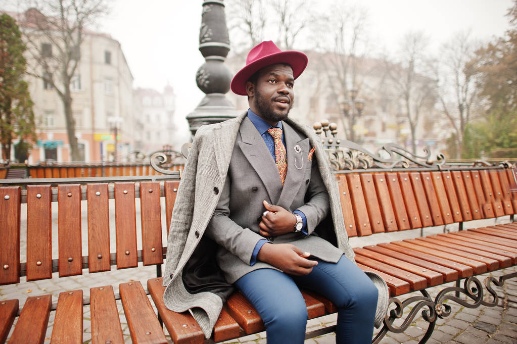 stylisches afrikanisch-amerikanisches Männermodel in grauem Mantel, Sakko-Krawatte und rotem Hut posierte an der Bank. - Foto, Bild