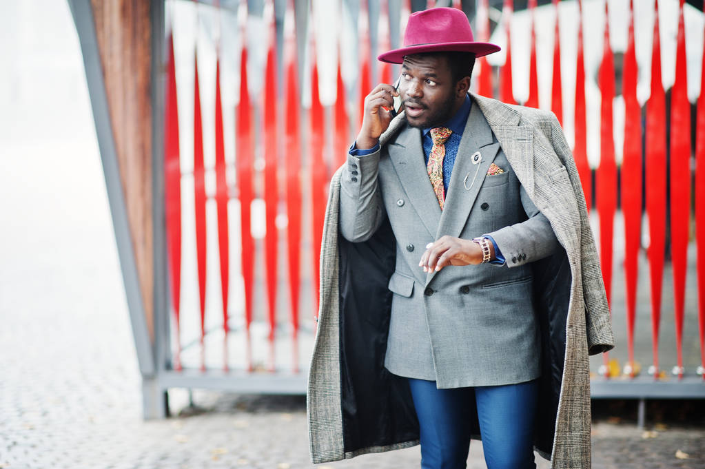 Stílusos afrikai amerikai férfi modell szürke kabát, kabát, nyakkendő és piros kalap, piros háttér kávézó, órák keresi és beszél a mobiltelefon szemben. - Fotó, kép