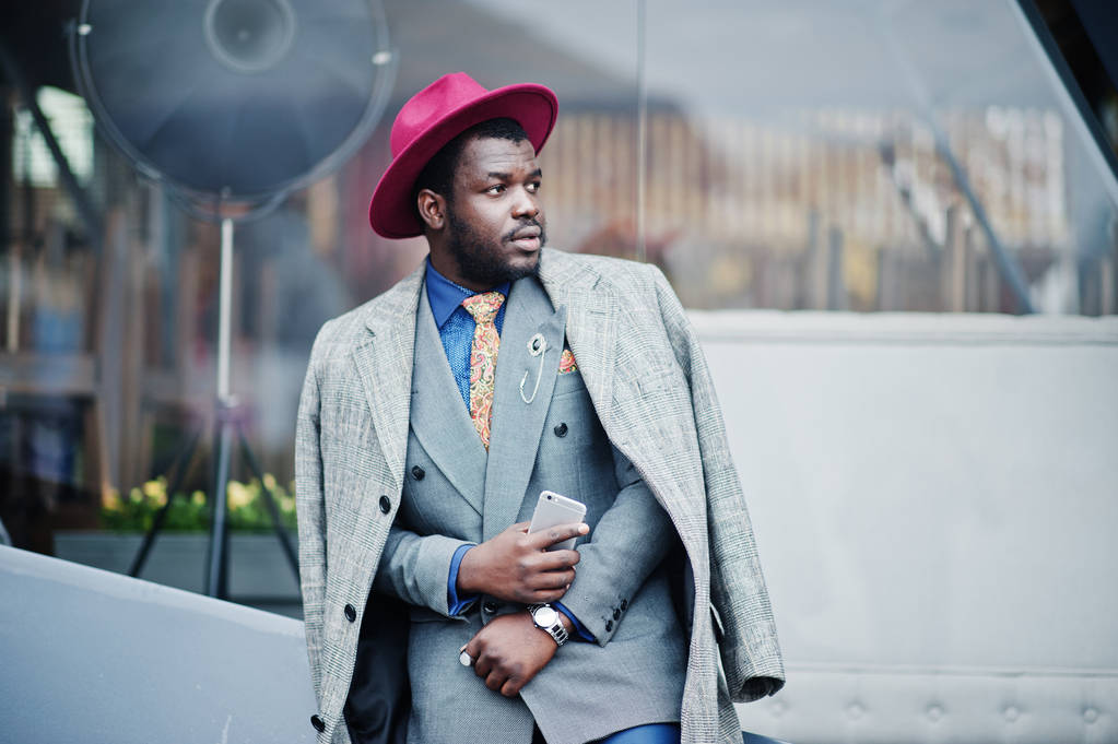 Elegante modelo de hombre afroamericano en abrigo gris, corbata de chaqueta y sombrero rojo con teléfono móvil en las manos
. - Foto, Imagen