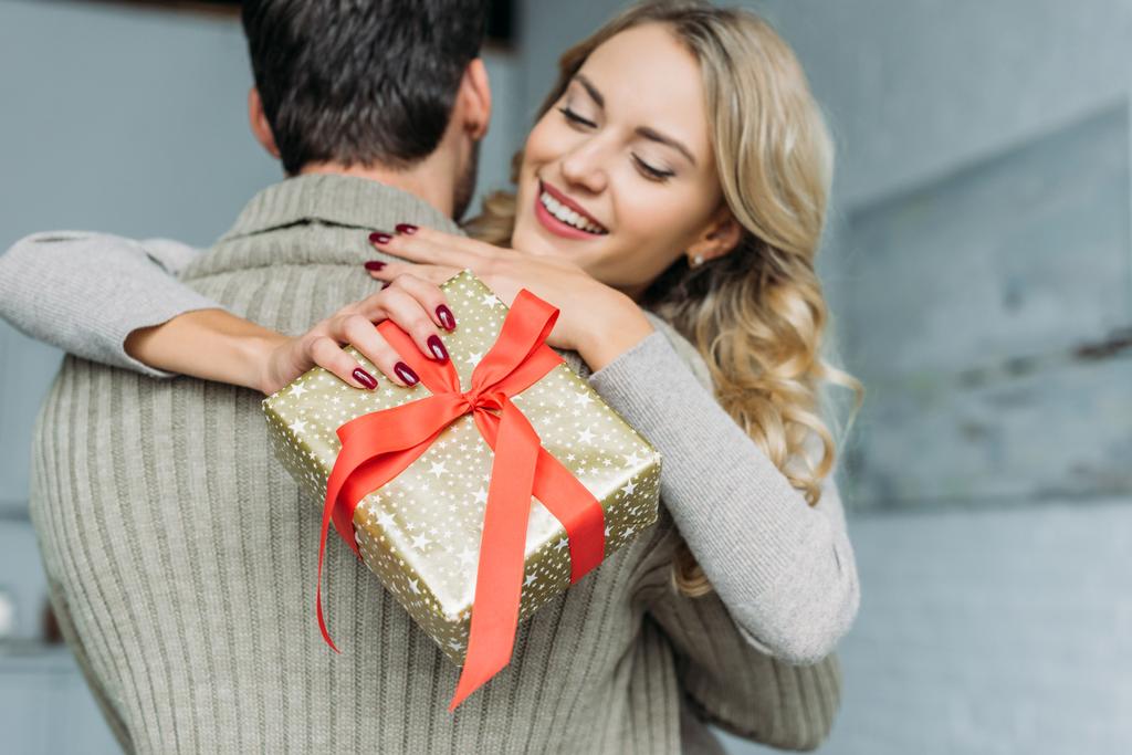beautiful young woman with gift box embracing her boyfriend at home - Photo, Image