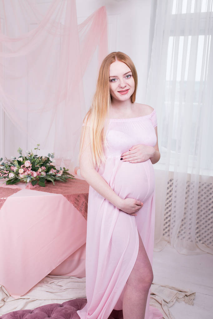 Młoda piękna kobieta w ciąży w różowej sukience stojącego obok okna w hom - Zdjęcie, obraz