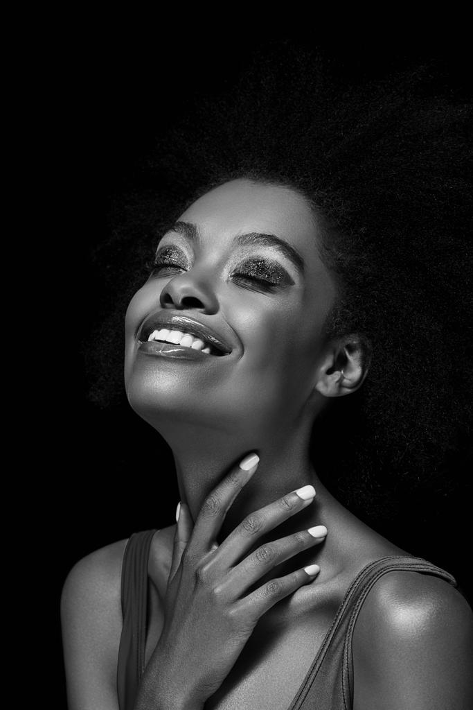 černo-bílé fotografie usměvavé afroamerické ženy izolované na černém - Fotografie, Obrázek